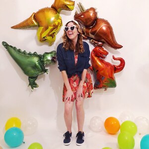Puede incluir: Una persona con gafas de sol blancas en forma de coraz&oacute;n, una camisa azul oscuro abotonada y un vestido rojo de flores con zapatillas azul oscuro. La persona est&aacute; rodeada de cuatro grandes globos de aluminio con forma de dinosaurio.