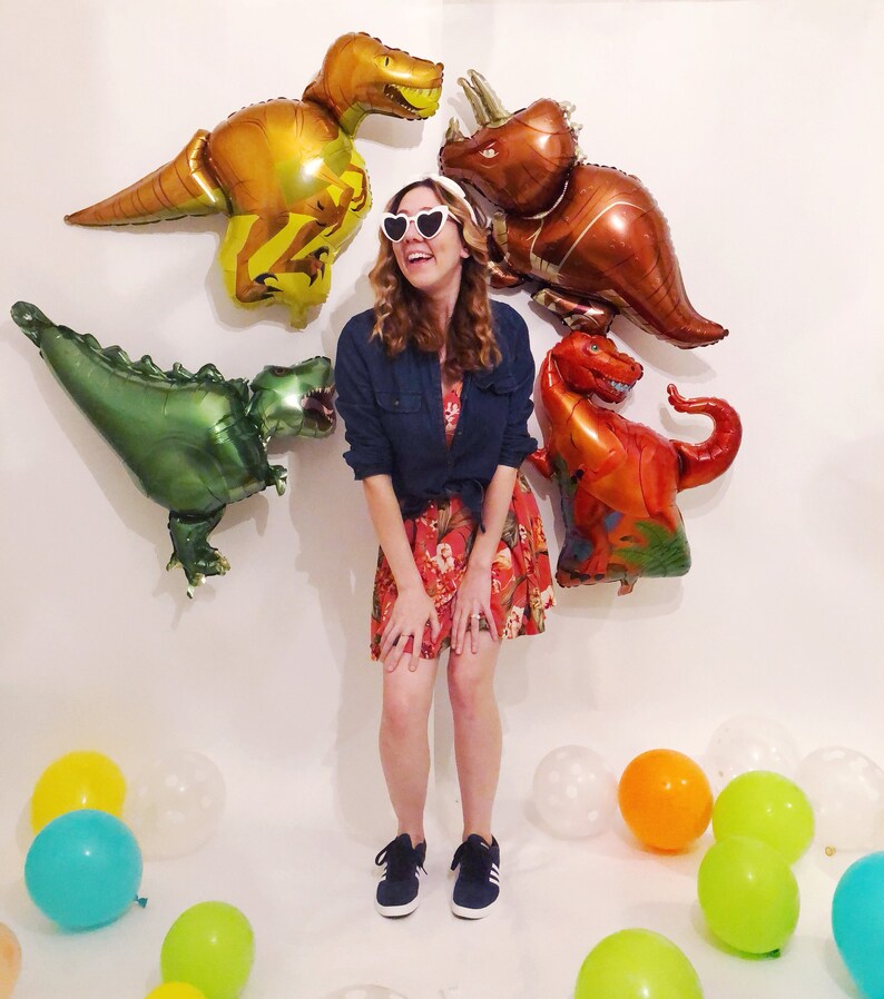 Puede incluir: Una persona con gafas de sol blancas en forma de coraz&oacute;n, una camisa azul oscuro abotonada y un vestido rojo de flores con zapatillas azul oscuro. La persona est&aacute; rodeada de cuatro grandes globos de aluminio con forma de dinosaurio.