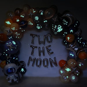 Puede incluir: Un arco de globos con dise&ntilde;os espaciales, incluyendo planetas, estrellas y cohetes, que brillan en la oscuridad. Globos plateados de aluminio deletrean "TWO THE MOON". Un globo de astronauta completa la decoraci&oacute;n.