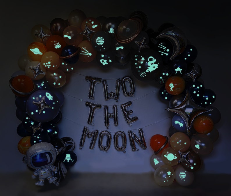 Puede incluir: Un arco de globos con dise&ntilde;os espaciales, incluyendo planetas, estrellas y cohetes, que brillan en la oscuridad. Globos plateados de aluminio deletrean "TWO THE MOON". Un globo de astronauta completa la decoraci&oacute;n.