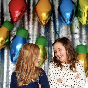 May include: A silver sequined backdrop decorated with multiple metallic foil balloons shaped like large Christmas light bulbs in red, blue, yellow, and green. Two children are positioned in front of the display, laughing and looking at each other.