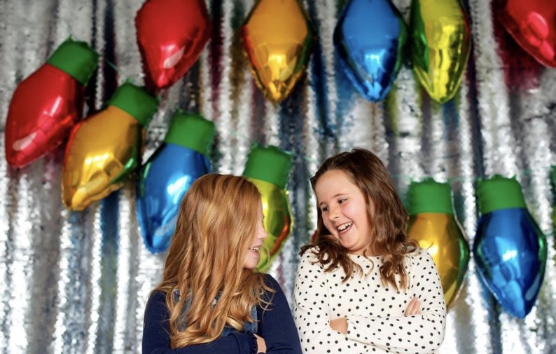 May include: A silver sequined backdrop decorated with multiple metallic foil balloons shaped like large Christmas light bulbs in red, blue, yellow, and green. Two children are positioned in front of the display, laughing and looking at each other.