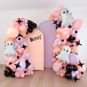 May include: A Halloween party backdrop with two arches made of pink, purple, and black balloons. The arches are decorated with ghost balloons and black star-shaped balloons. The text "BOO!" is written on a pink arch.