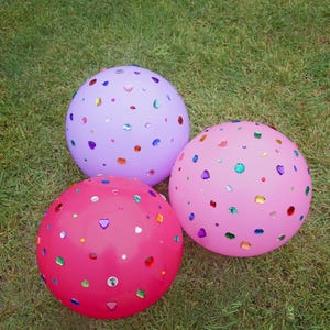 May include: Three round balloons in shades of light purple, bright pink, and hot pink, each decorated with an assortment of small, colorful, reflective plastic gems in various shapes including hearts, circles, and squares scattered across the surface.