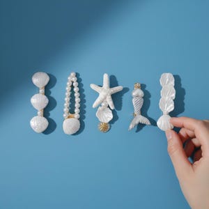 May include: A collection of decorative hair clips and barrettes with a sea life theme. The clips feature white seashells, pearls, and a mermaid design. The background is a solid blue.