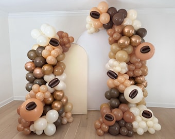 May include: A balloon arch and column in shades of brown, beige, and white, decorated with coffee bean-shaped balloons. The arrangement is set against a neutral backdrop, creating a festive and thematic display.