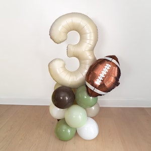 May include: A balloon arrangement featuring a large, cream-colored number "3" balloon, a brown football-shaped balloon, and a cluster of smaller balloons in shades of brown, green, and white. The balloons are arranged on a wooden floor.