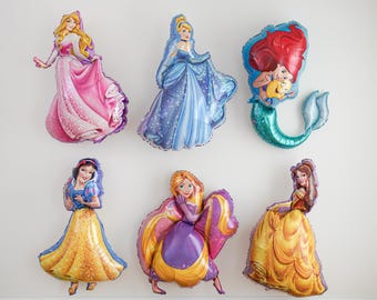 May include: A collection of six foil balloons shaped like various animated princesses. The balloons feature characters in pink, blue, yellow, and purple gowns, along with a mermaid character with red hair and a green tail, all with glossy finishes.