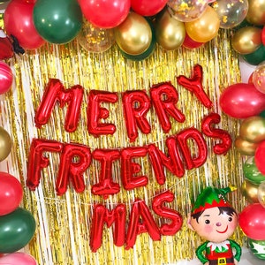 May include: A festive decoration featuring a gold tinsel fringe backdrop framed by red, green, and gold balloons. Red foil letter balloons spell out MERRY FRIENDS MAS. A small elf-shaped foil balloon stands at the bottom right corner.