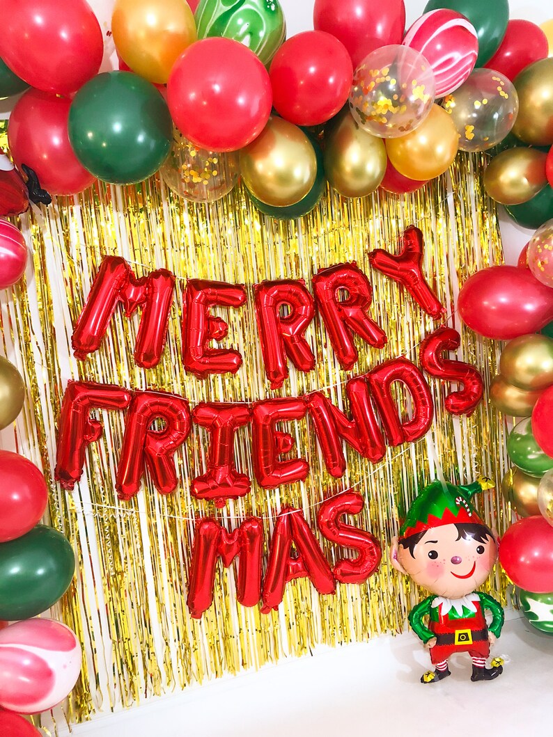 May include: A festive decoration featuring a gold tinsel fringe backdrop framed by red, green, and gold balloons. Red foil letter balloons spell out MERRY FRIENDS MAS. A small elf-shaped foil balloon stands at the bottom right corner.
