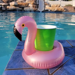 K&ouml;nnte beinhalten: Aufblasbarer rosa Flamingo-Poolschwimmer mit einem gr&uuml;nen Becherhalter. Der Flamingo hat einen schwarzen Schnabel und ein gelbes Auge. Der Schwimmer befindet sich am Rand eines blau gefliesten Pools.