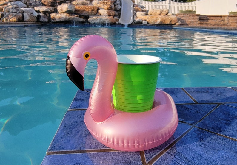 K&ouml;nnte beinhalten: Aufblasbarer rosa Flamingo-Poolschwimmer mit einem gr&uuml;nen Becherhalter. Der Flamingo hat einen schwarzen Schnabel und ein gelbes Auge. Der Schwimmer befindet sich am Rand eines blau gefliesten Pools.