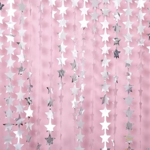 May include: Silver star-shaped garlands hanging against a soft pink background. The metallic stars are strung together, creating a shimmering, festive decoration. Ideal for parties, celebrations, or adding a touch of sparkle to any event.
