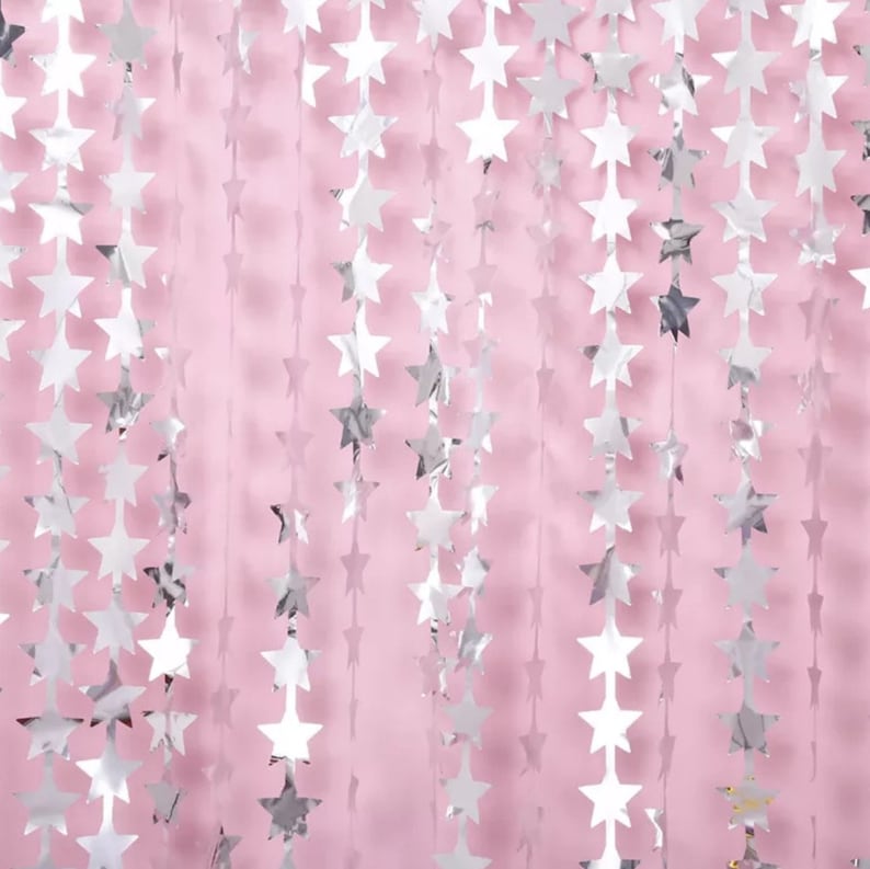 May include: Silver star-shaped garlands hanging against a soft pink background. The metallic stars are strung together, creating a shimmering, festive decoration. Ideal for parties, celebrations, or adding a touch of sparkle to any event.