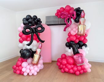 May include: Two balloon structures in pink, black, and white, decorated with bow and lipstick-shaped balloons. One structure has a nail polish balloon. The structures are set against a pink arch and a black and white striped backdrop.