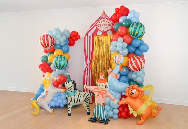 K&ouml;nnte beinhalten: Eine Zirkus-Themen-Ballonbogen mit roten, blauen, gr&uuml;nen und gelben Ballons. Aufblasbare Zirkusfiguren wie Zebra, L&ouml;we, Clown und Seehund. Eine Zirkuszelt-Kulisse mit goldenen Fransen.
