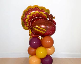 May include: A Thanksgiving-themed balloon arrangement featuring a large, detailed turkey balloon in shades of red, brown, and gold, atop a column of colorful balloons in orange, yellow, and purple. Perfect for festive decorations.