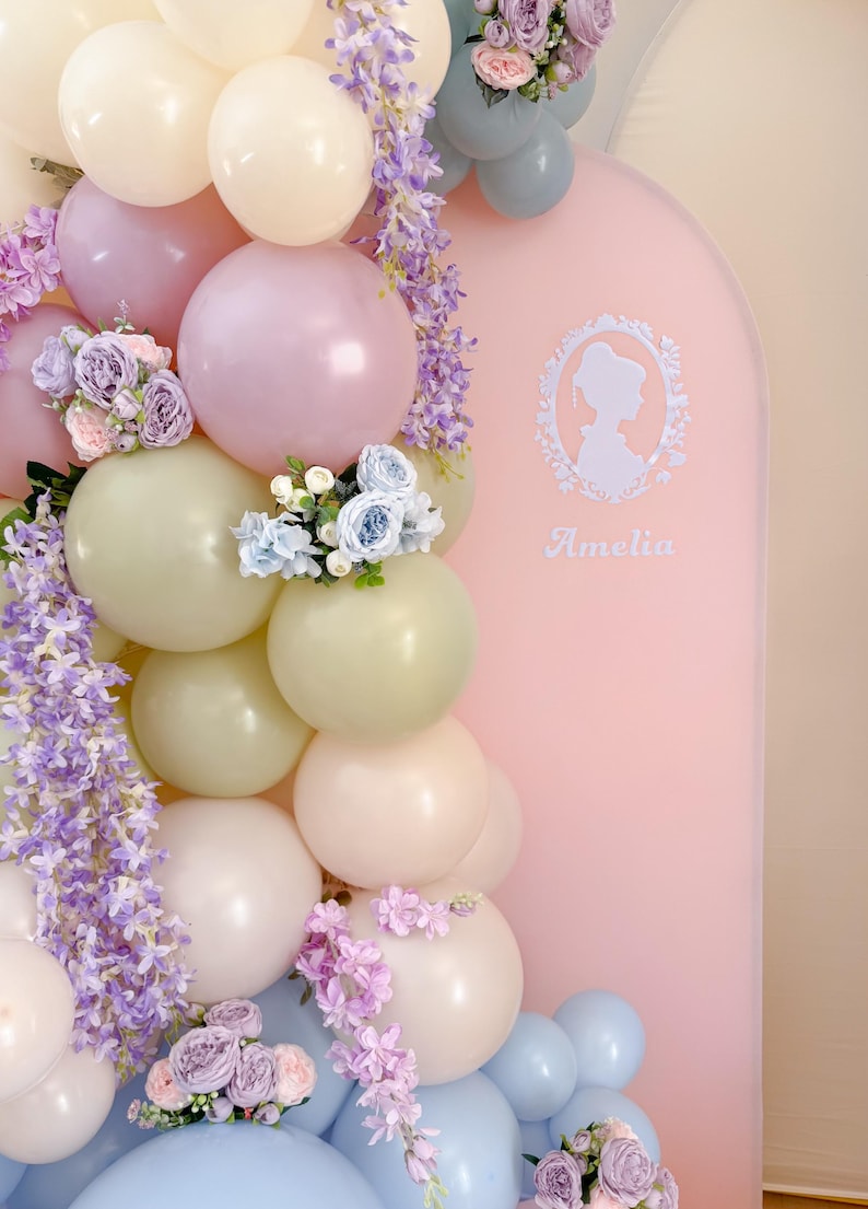 May include: A pastel balloon arch with a pink backdrop, decorated with faux flowers and wisteria. The balloons are in shades of white, pink, and green. The backdrop features a silhouette and the name "Amelia".