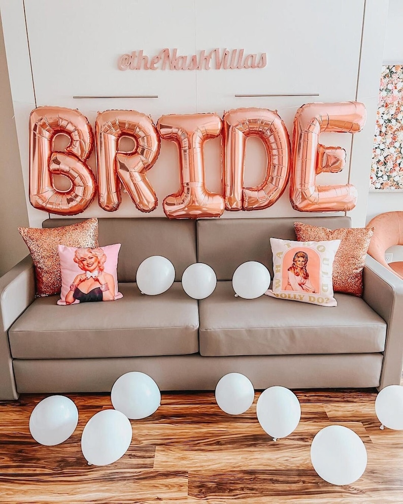 May include: A rose gold foil balloon display spelling out the word "BRIDE" is suspended above a gray sofa. Decorative pillows with sequin and portrait designs are arranged on the sofa, along with white balloons. The text "@theNashVillas" is above the balloons.