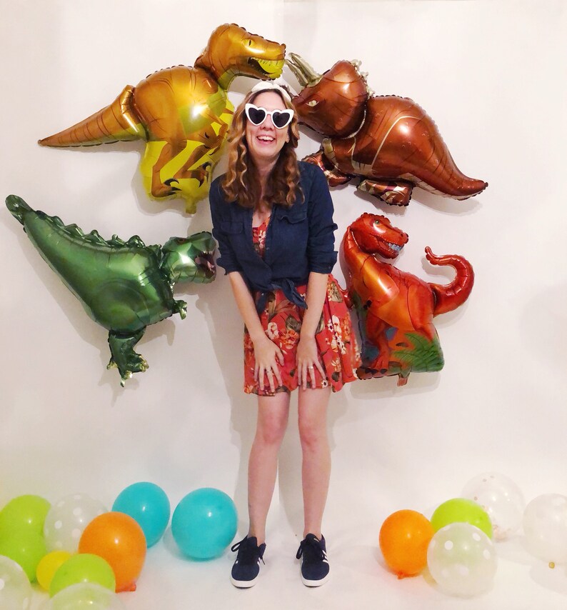 Puede incluir: Una persona con una camisa azul oscuro, un vestido rojo con estampado floral y gafas de sol blancas en forma de coraz&oacute;n, rodeada de cuatro globos de aluminio con forma de dinosaurio y varios globos redondos naranjas, verdes y azules.