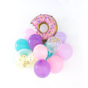 May include: A cluster of balloons featuring a large, pink, donut-shaped foil balloon with colorful sprinkles and a bite mark. The arrangement includes solid-colored latex balloons in shades of purple, pink, and teal, along with clear balloons filled with gold confetti.