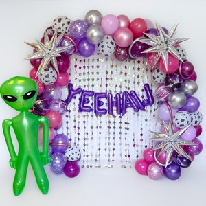 May include: A purple and silver balloon arch with the word "Yeehaw" spelled out in purple letters. The arch is decorated with stars and is surrounded by a variety of balloons in different colors and patterns. A green alien balloon is in the foreground.