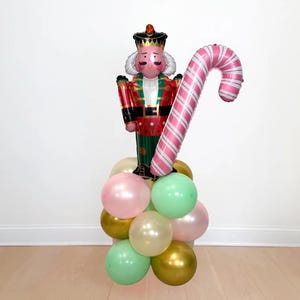 May include: A festive balloon arrangement featuring a nutcracker soldier and a pink and white striped candy cane. The base consists of pastel pink, green, gold, and cream-colored balloons, creating a cheerful holiday display.