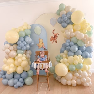 May include: A baby sits in a high chair decorated with a banner that says "1" in front of a balloon arch made of blue, cream, and green balloons. The balloon arch is in the shape of an archway.