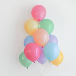 May include: A cluster of eleven round latex balloons in a variety of pastel colors including pink, yellow, mint green, peach, light blue, and lavender. The balloons are arranged in a vertical, tree-like formation with white ribbons tied at the base.