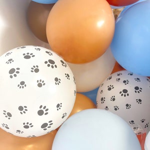 Puede incluir: Un conjunto de globos coloridos en tonos de azul, naranja, marr&oacute;n y blanco. Algunos globos son lisos, mientras que otros presentan un patr&oacute;n de peque&ntilde;as huellas de patas negras. Los globos est&aacute;n agrupados.