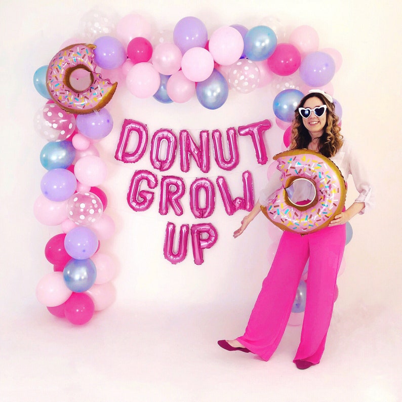 Pode incluir: Um arco de bal&otilde;es cor-de-rosa, roxos e azuis com dois bal&otilde;es de alum&iacute;nio grandes em forma de donut. O texto DONUT GROW UP &eacute; formado por bal&otilde;es de letras cor-de-rosa. Uma pessoa usa &oacute;culos de sol brancos em forma de cora&ccedil;&atilde;o e cal&ccedil;as cor-de-rosa.