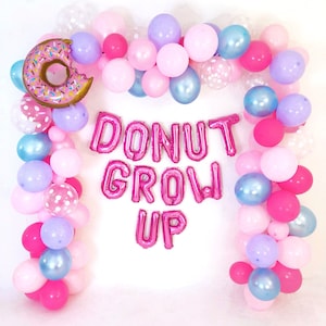 Pode incluir: Um arco de bal&otilde;es com bal&otilde;es cor-de-rosa, azul-claros, roxos e brancos com bolinhas, com um bal&atilde;o de alum&iacute;nio em forma de donut no canto superior esquerdo. O centro exibe bal&otilde;es de letras cor-de-rosa que formam DONUT GROW UP.