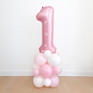 May include: A pink number one foil balloon with white cross accents, atop a cluster of pink and white balloons. The balloon arrangement is a festive decoration for a first birthday celebration.