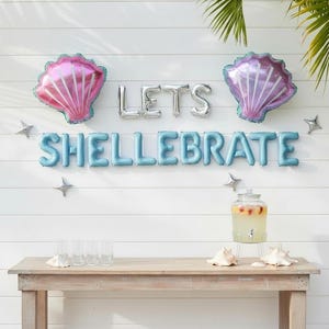 May include: A festive outdoor scene with a wooden table and white wall backdrop. The wall is decorated with seashell-shaped balloons in pink and purple, silver star balloons, and the phrase "LETS SHELLEBRATE" in silver and blue balloons. A glass beverage dispenser and seashells are on the table.