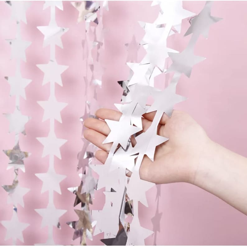 May include: Silver star-shaped garland hanging against a pink background. The garland is made of reflective material, creating a shimmering effect. The stars are connected in a chain, suitable for party decorations.