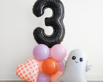 May include: A balloon arrangement featuring a large black foil number 3 balloon atop a cluster of round lavender, pink, and orange balloons. A heart-shaped balloon with an orange and white gingham pattern and a white ghost balloon are attached.