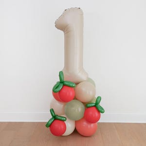 May include: A beige number one balloon is the centerpiece of a balloon arrangement. The base is composed of various balloons in shades of red, green, and beige. The arrangement is suitable for a first birthday celebration.