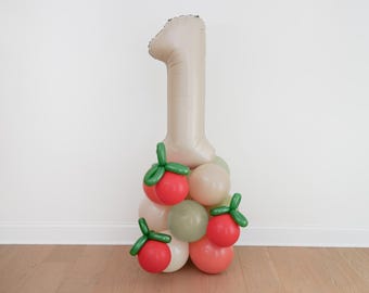 May include: A beige number one balloon is the centerpiece of a balloon arrangement. The base is composed of various balloons in shades of red, white, and green, some shaped like strawberries. This festive decoration is suitable for a first birthday celebration.