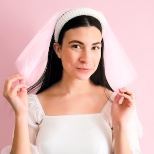 May include: A white bridal headband with pearl accents and a pink tulle veil. The headband is worn on a person's head.