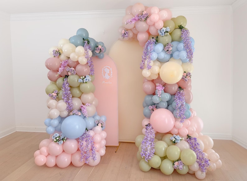 May include: A decorative balloon display in pastel colours, featuring pink, blue, green, and cream balloons. The arrangement is decorated with purple wisteria and small floral accents. A pink arch with a silhouette is in the background.
