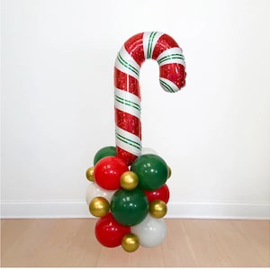 May include: A large foil candy cane balloon with red, white, and green stripes, standing on a base made of a cluster of smaller round balloons in red, dark green, white, and metallic gold colors, creating a festive holiday decoration.
