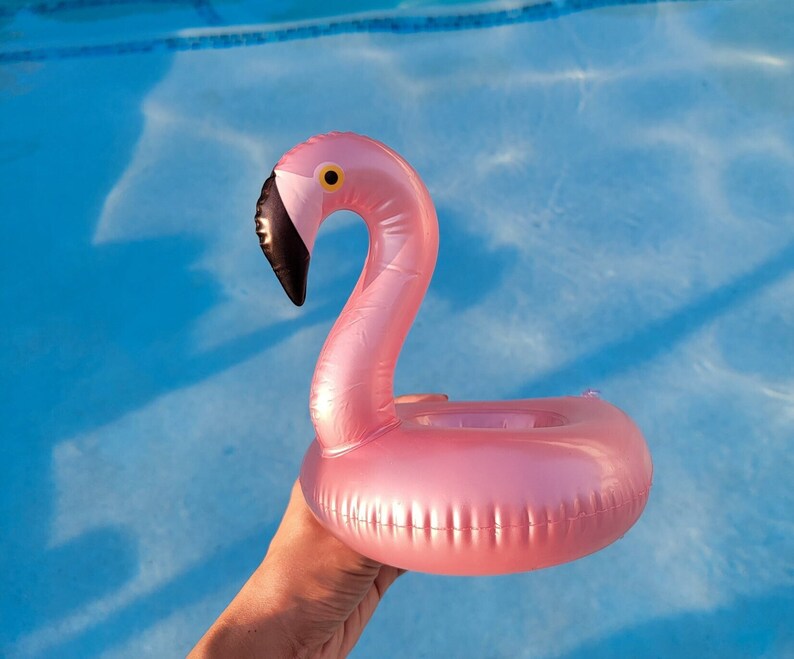 K&ouml;nnte beinhalten: Ein aufblasbarer Getr&auml;nkehalter in Form eines rosa Flamingos mit schwarzem Schnabel und gelbem Auge, vor blauem Wasser. Der Flamingo ist zum Schwimmen im Pool und zum Halten eines Getr&auml;nks konzipiert.