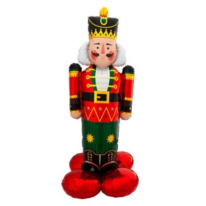 May include: A nutcracker-shaped foil balloon featuring a red, green, and black uniform with gold accents, a tall black hat with a green gem, white hair, and a painted face. The balloon stands on a base made of four round red foil balloons.