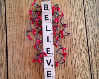 May include: A wooden sign spelling "BELIEVE" using Scrabble tiles. The letters are arranged vertically and decorated with red berry sprigs and a woven loop for hanging. The sign is set against a wooden background.