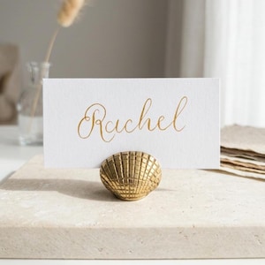 Può includere: Un portacarte a forma di conchiglia dorata con un biglietto bianco con la scritta "Rachel" in oro. Il supporto è su una superficie di pietra chiara, suggerendo un tema costiero.