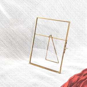 May include: A rectangular, gold-coloured metal picture frame with a clear glass pane. The frame has a hinged stand on the back for tabletop display. The frame is empty and ready to hold a photograph or artwork.