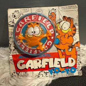 May include: A vintage Garfield the cat yo-yo toy. The yo-yo features a 3D orange Garfield figure in a blue and red circular frame. The packaging includes comic strip illustrations and the words "Garfield Yo-Yo".