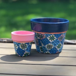 Op de afbeelding: Twee beschilderde terracotta bloempotten, een roze en een blauwe, met een witte achtergrond en blauwe, groene en roze geometrische patronen.