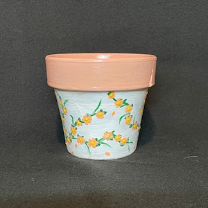 May include: A small terracotta flower pot with a white base and a pink rim. The pot is decorated with a hand-painted design of green vines and orange flowers.