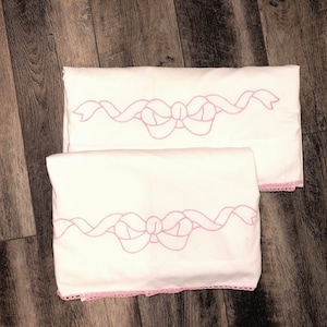 Puede incluir: Dos fundas de almohada blancas con diseños de cinta y lazo bordados en rosa. Las fundas de almohada tienen un delicado borde de encaje.